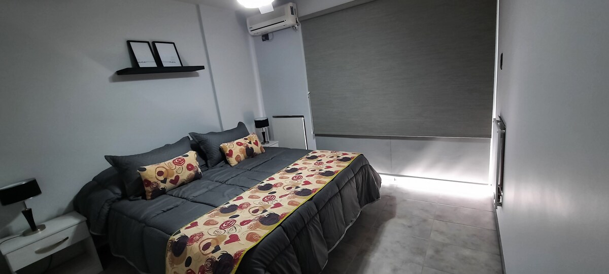 A spacious bedroom features a large bed covered with a patterned blanket. Two decorative pillows add a pop of color. Shelves with framed items are mounted above the bed, while a window is partially covered by a roller shade, allowing for controlled light.