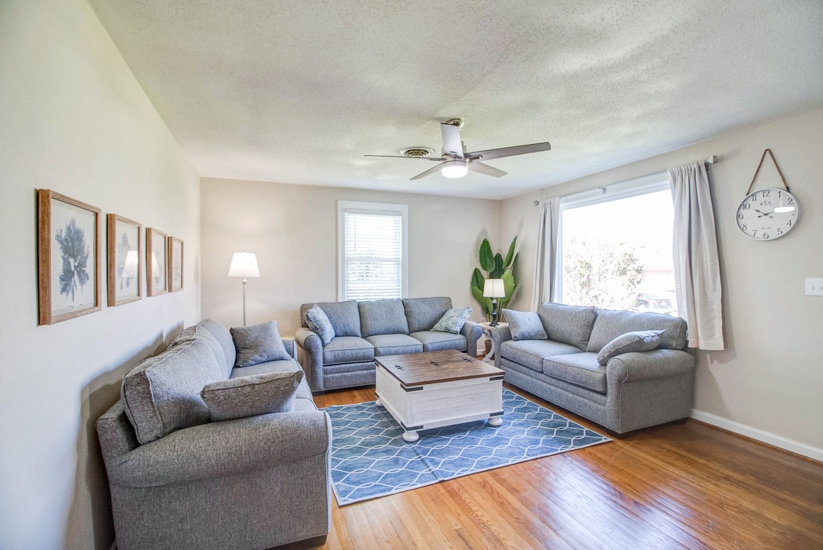 A spacious living room features multiple soft gray sofas arranged around a central coffee table. A patterned blue area rug covers the hardwood floor. Natural light enters through a large window, complemented by light curtains. Decorative wall art and a clock add character to the space.