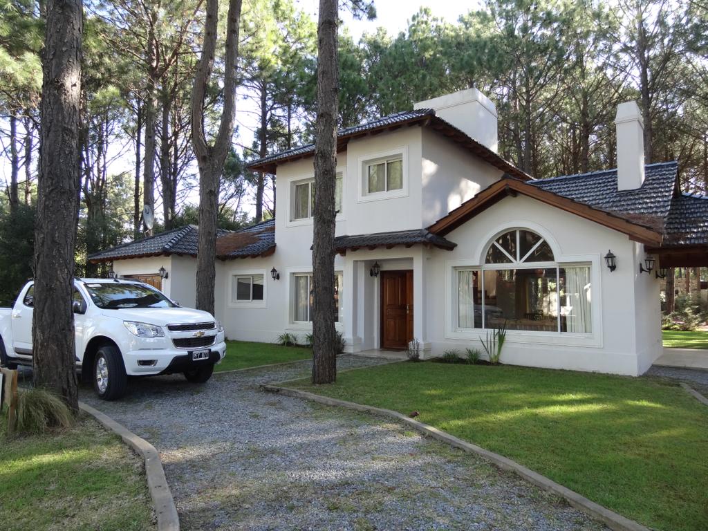 A two-story chalet is surrounded by tall trees, featuring a well-maintained lawn. The exterior has a combination of white walls and a wooden roof. A large arched window adds character, while a gravel path leads up to the entrance, alongside a parked vehicle.