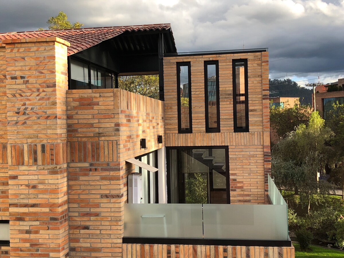 The exterior of a modern building showcases a combination of textured brick and sleek glass. Large windows allow natural light to enter, while a balcony extends from the structure, providing an outdoor space. The surrounding landscape features greenery and city views.