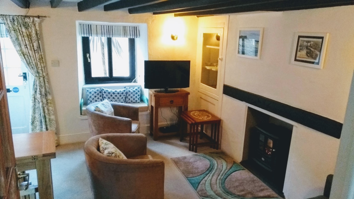 A cozy living room features a low ceiling with exposed wooden beams and light walls. Comfortable tub chairs are positioned around a small coffee table. A flat screen TV is placed on a wooden stand, with a quaint electric fireplace nearby. A window seat offers views of the outside.
