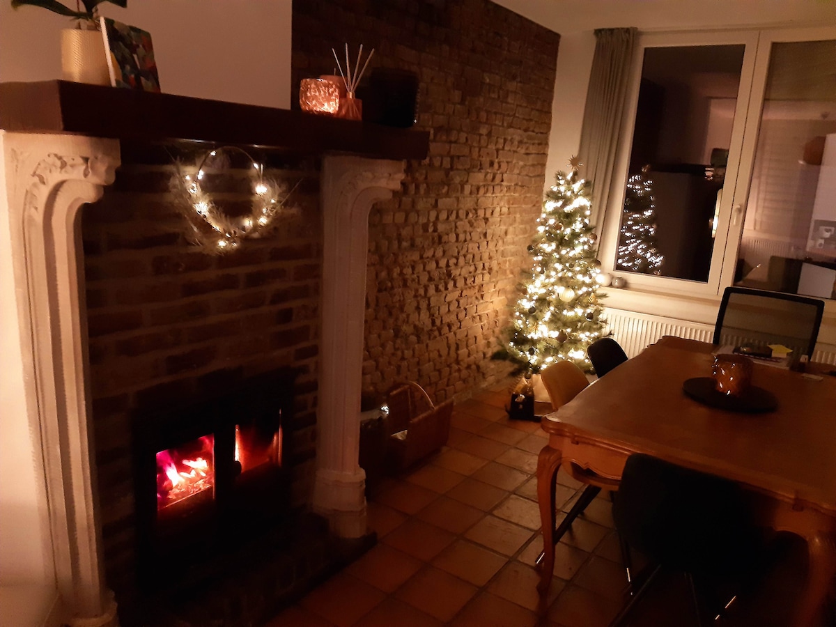 A cozy living area features a warm fireplace with glowing embers and a rustic stone wall. A small Christmas tree is adorned with lights, while a wooden dining table is set with a centerpiece. Soft lighting enhances the inviting atmosphere.