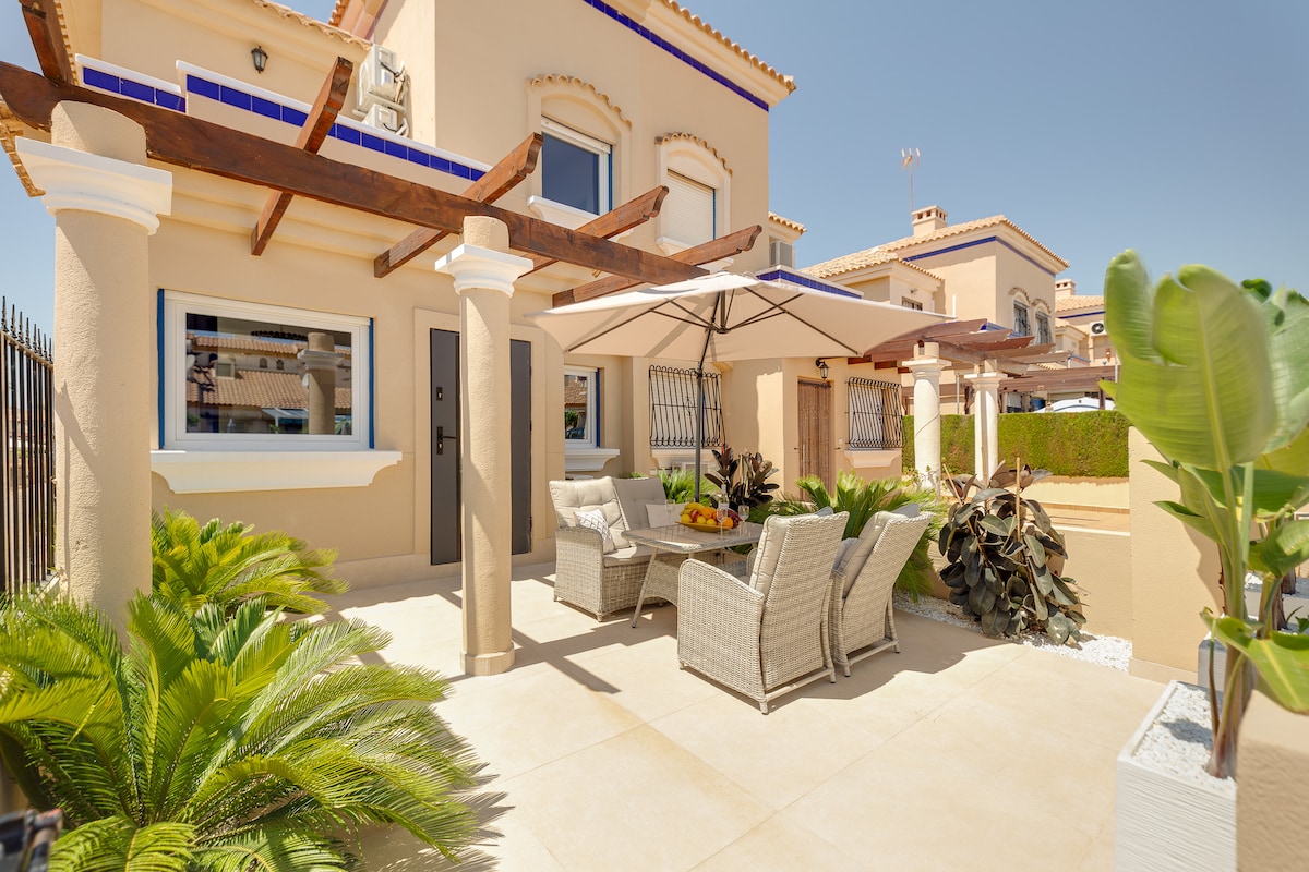 A shaded patio area features comfortable wicker furniture, surrounded by lush greenery. Decorative plants are positioned alongside the seating arrangement, with a stone pathway leading to the entrance. Sunlight illuminates the space, highlighting the home's architectural details.