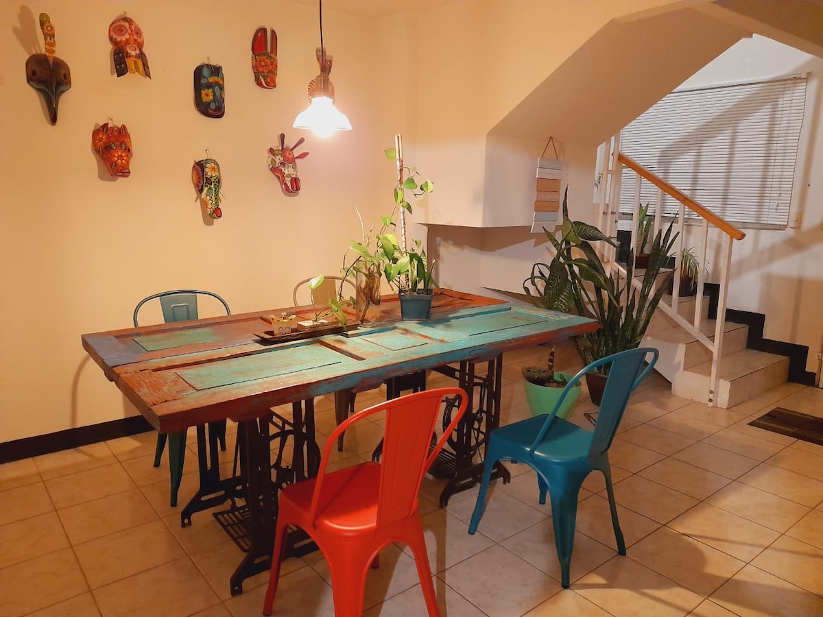 A spacious dining area features a large, colorful table surrounded by metal chairs in vibrant hues of red and blue. Decorative masks are mounted on the wall, while potted plants add a touch of greenery. Natural light filters through a nearby window.