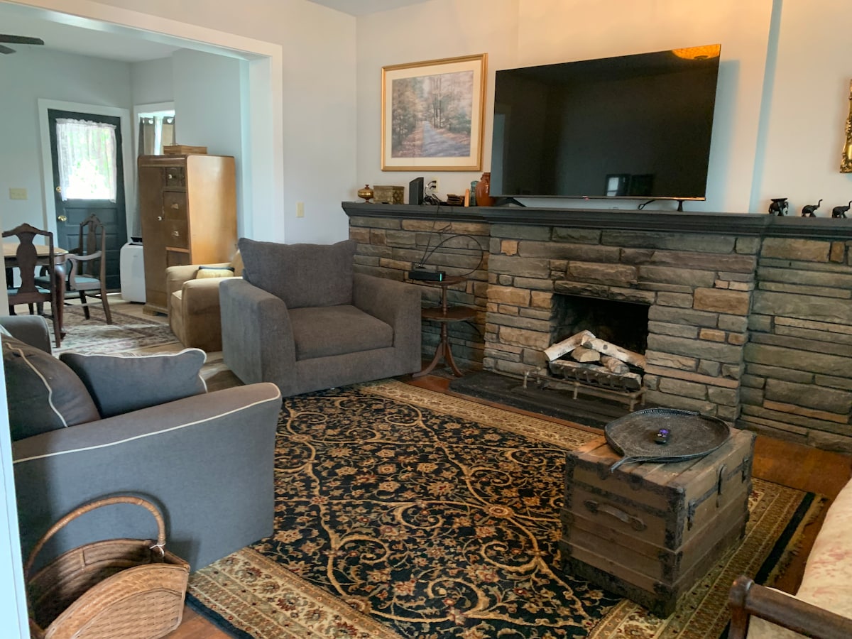 A cozy living area features a stone fireplace with decorative elements, a large television mounted above it, and comfortable seating including a spacious armchair and a sofa. A patterned area rug covers the hardwood floor, with a wooden trunk and a woven basket adding to the decor.
