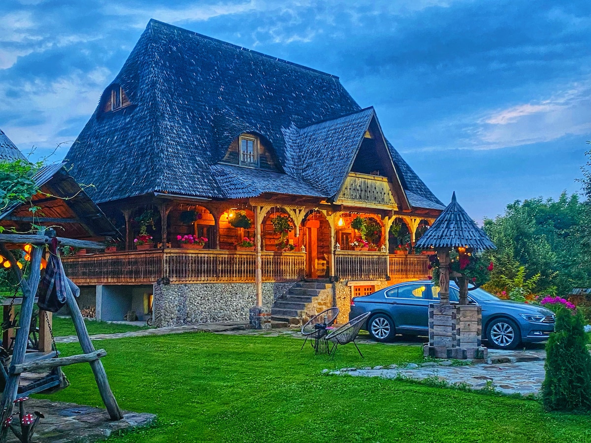 A traditional wooden house features an intricate roof and is illuminated by warm outdoor lights. The property includes a well-maintained lawn with seating areas and a decorative wooden well, creating a peaceful outdoor space for relaxation.