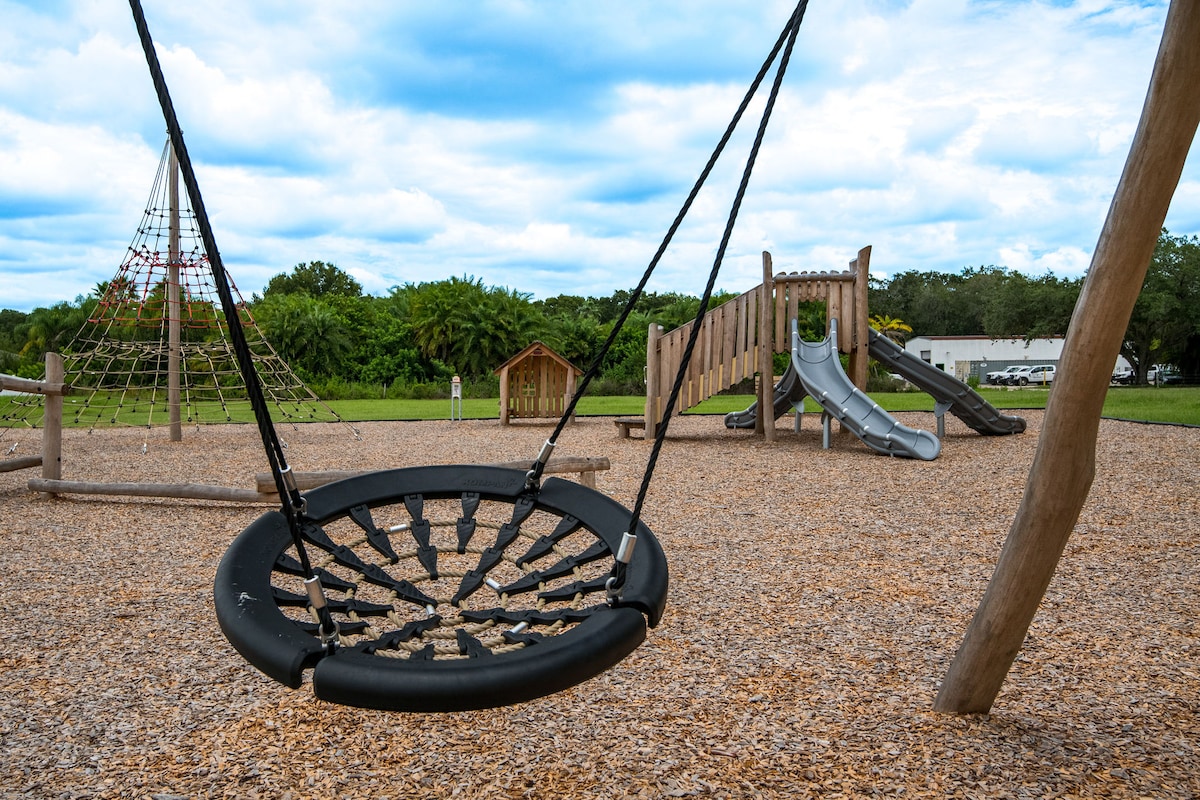 A children's playground is featured, showcasing a large swing, climbing structure, and a slide, all situated on a bed of wood chips. Lush greenery surrounds the area, with a playground structure constructed from natural materials, providing a welcoming space for outdoor play.