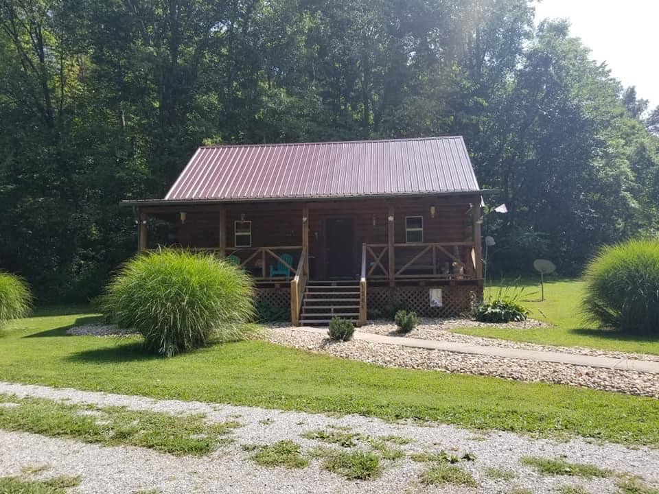 A rustic log cabin features a red metal roof and a welcoming porch with wooden railings. Lush greenery surrounds the cabin, including tall ornamental grasses and neatly arranged stones. A gravel pathway leads up to the entrance, providing an inviting approach to the home.