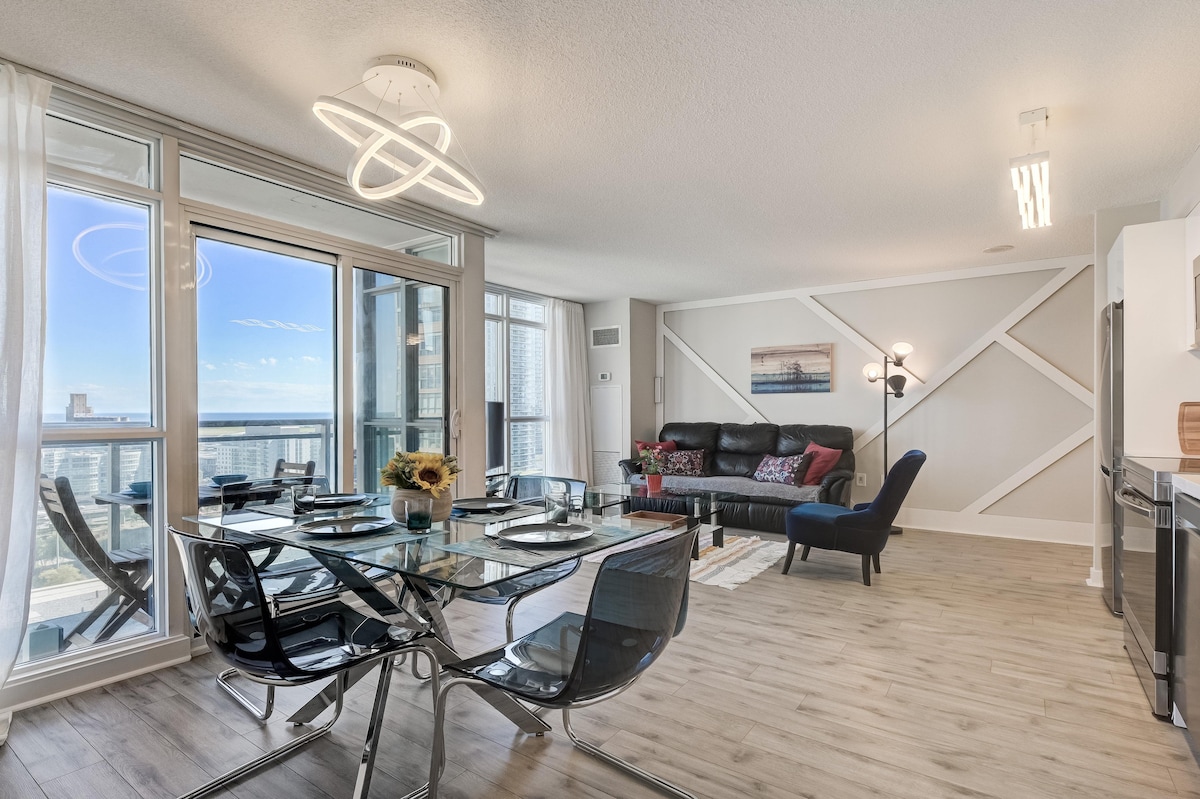 The open-plan living area showcases floor-to-ceiling windows offering views of the city skyline and lake. A glass dining table with black chairs is set for dining, while a sofa with decorative pillows and a stylish accent chair complete the space. Modern light fixtures enhance the ambiance.