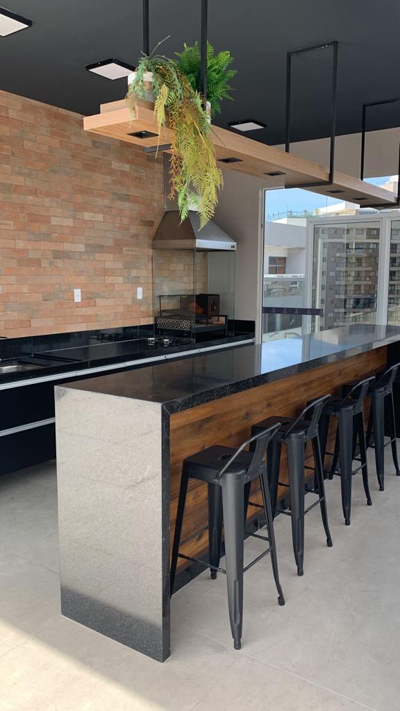 A modern kitchen features an island with high black stools, complemented by a sleek granite countertop. Natural wood accents and a brick wall impart warmth. A stainless steel hood is mounted above the cooking area, while large windows allow for ample natural light and views of the city.