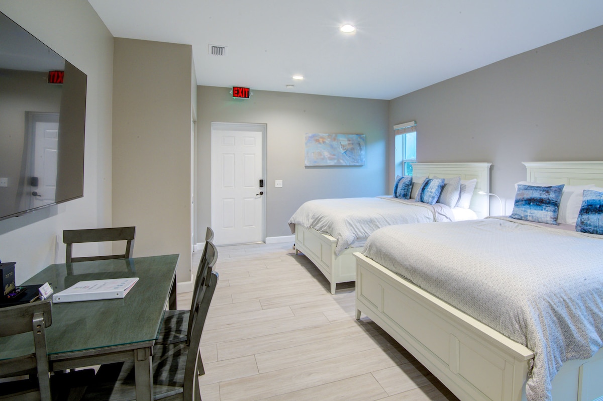 A spacious studio is depicted featuring two queen beds with light bedding and decorative pillows. A flat-screen television is mounted on the wall in front of the beds. A small dining table and chairs are positioned nearby, and natural light enters through a window.