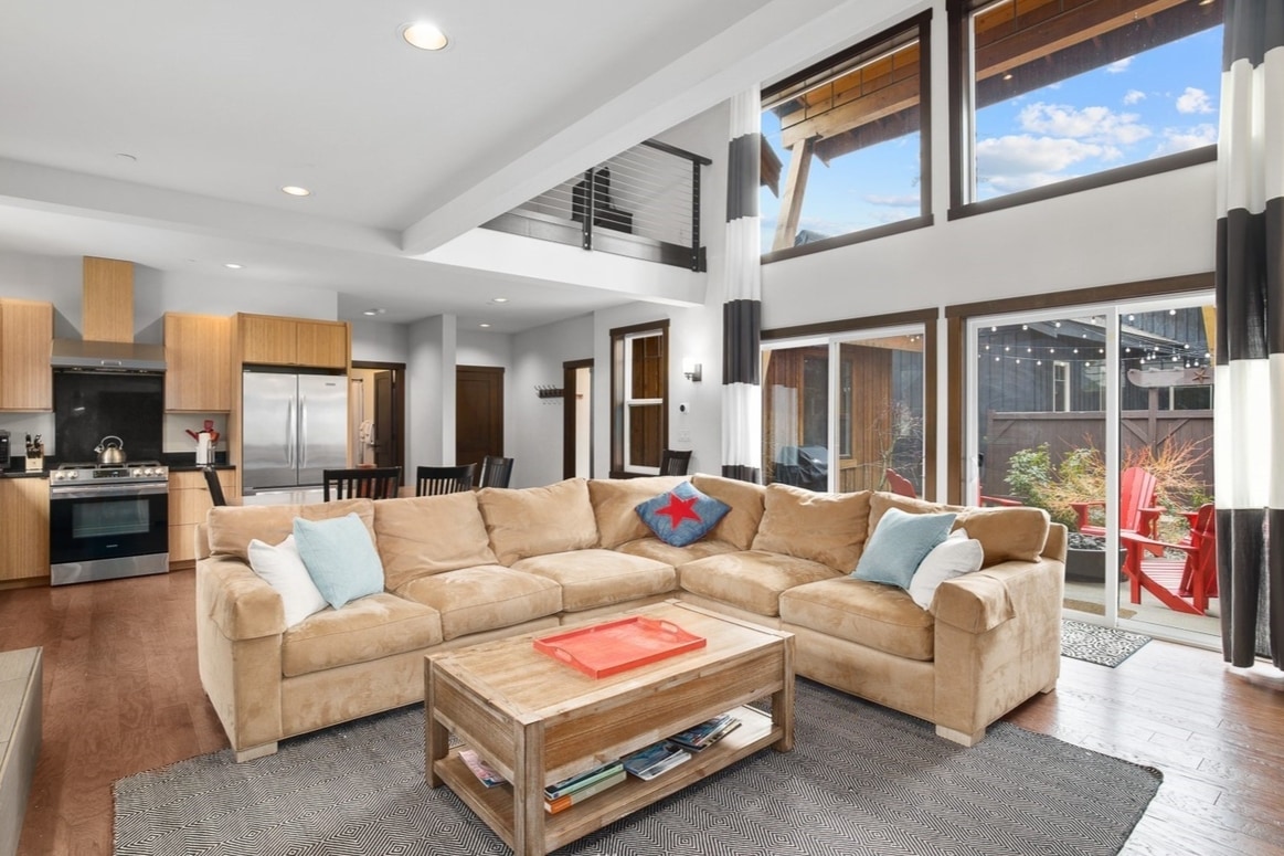 A spacious living area features a comfortable sectional couch adorned with colorful accent pillows. A wooden coffee table sits in front, while large windows allow natural light to fill the room. The contemporary kitchen is visible in the background, contributing to the open layout.