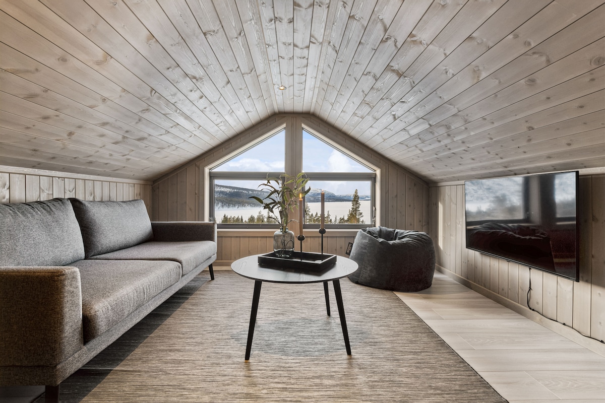 A cozy loft area features a spacious gray sofa, complemented by a circular coffee table. Two large windows frame picturesque views of the surrounding landscape, while a television is mounted on the wall. Soft lighting highlights the wooden ceiling and flooring.