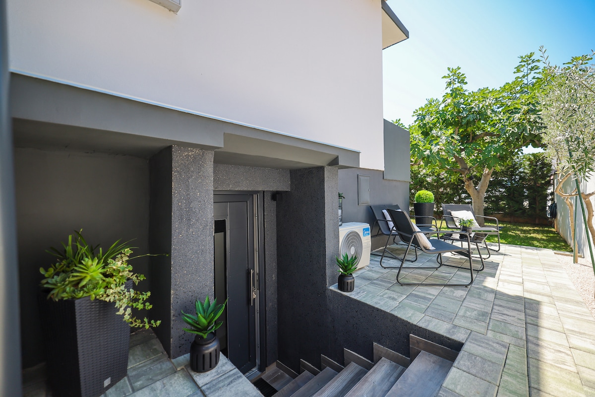 A modern entrance leads to the apartment, with a staircase flanked by potted plants. An outdoor seating area is visible, featuring two lounge chairs and surrounded by greenery. Sunlight brightens the space, highlighting the clean lines and contemporary design.