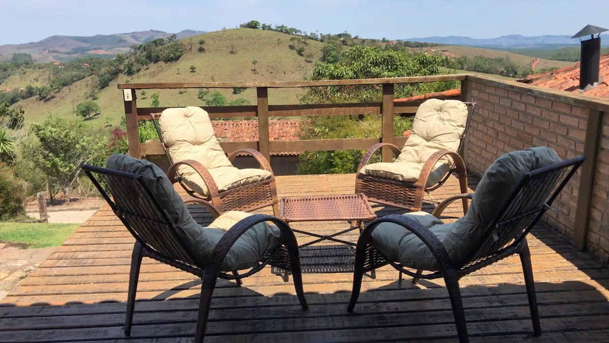 A spacious wooden deck features two comfortable armchairs with soft cushions and a small table between them. The area offers unobstructed views of rolling hills and greenery, creating a serene outdoor space for relaxation.