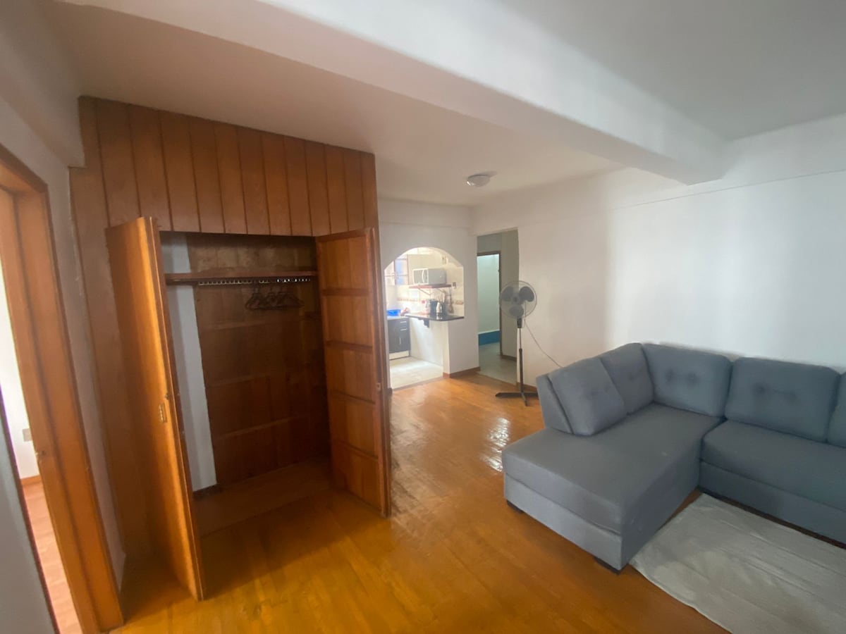 A cozy living space features a gray sectional sofa positioned near a wooden closet. Natural light filters through the room, highlighting the warm wooden floors. The open layout leads to a kitchenette area, with a glimpse of appliances in the background.