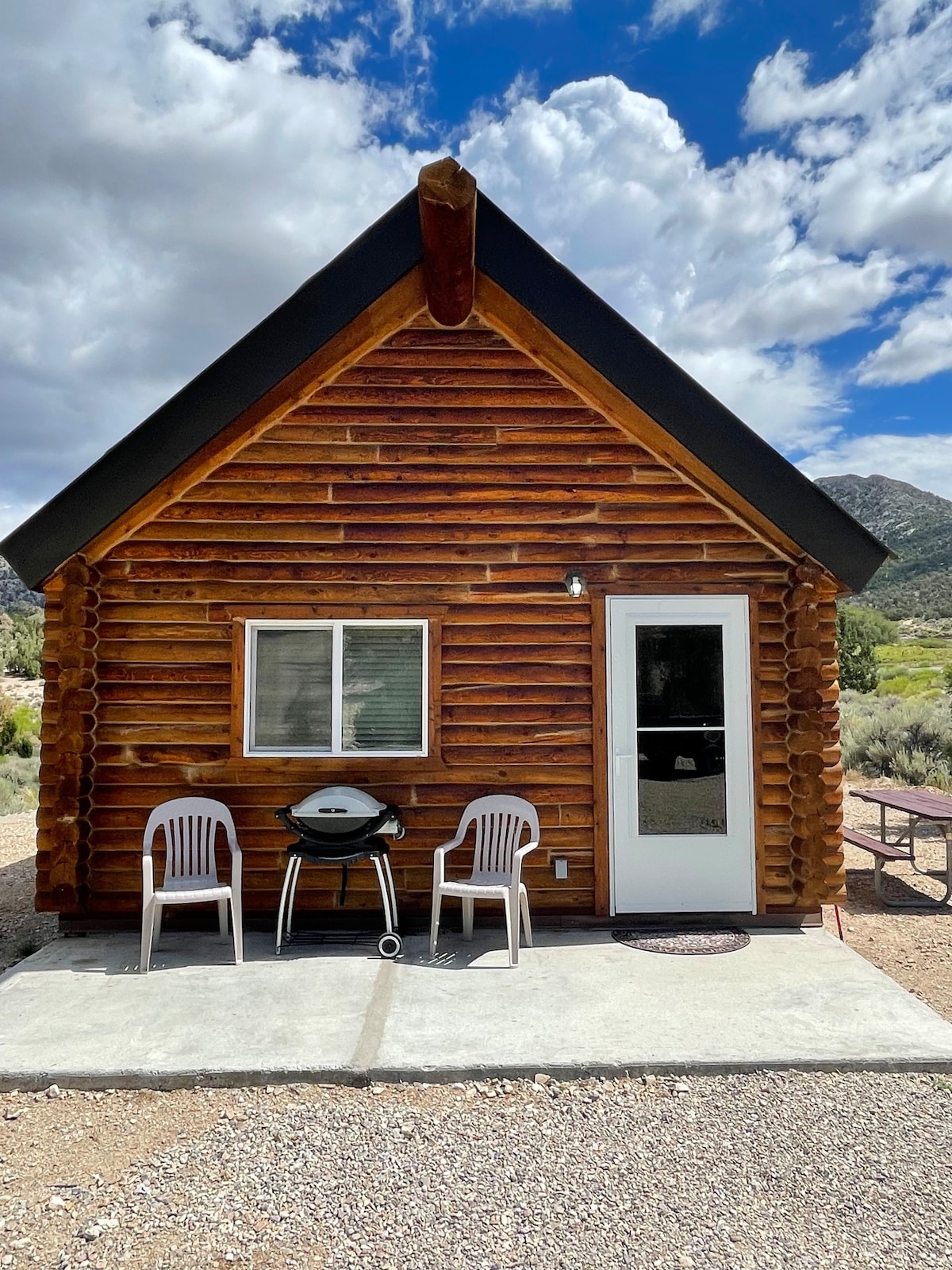 The exterior of a cozy cabin is shown, featuring a log construction and a sloped roof. A small patio area includes two white plastic chairs and a grill, while a picnic table is visible nearby. The surrounding landscape displays greenery and distant mountains under a partly cloudy sky.