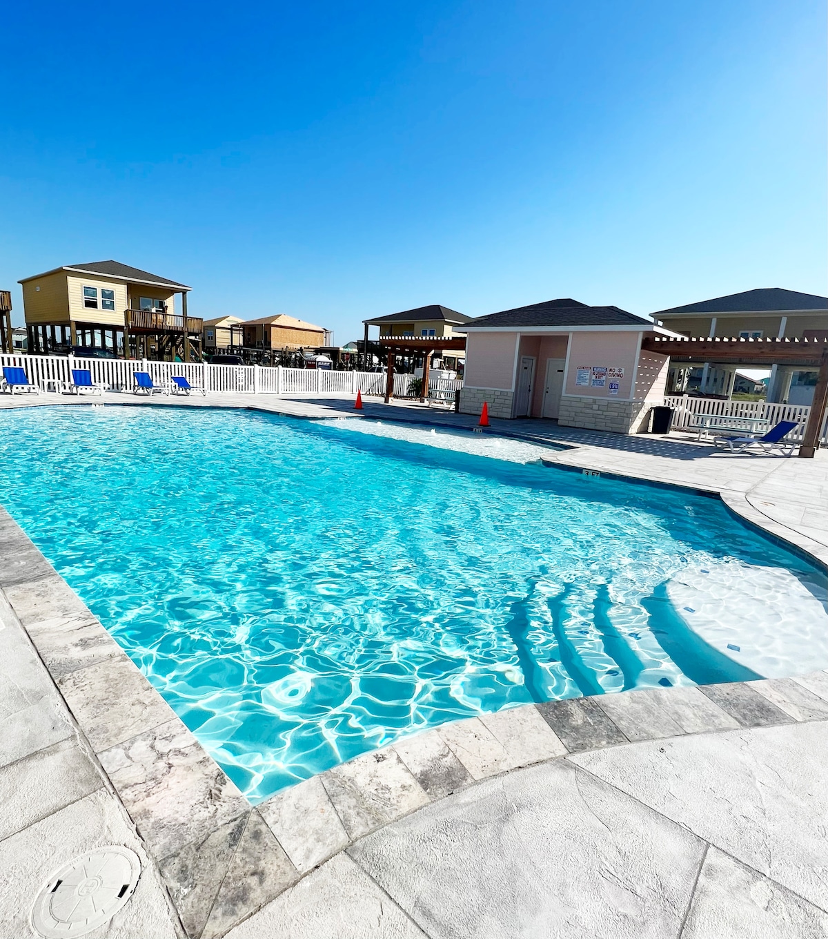 A community pool is framed by a white fence and lounge chairs under a clear blue sky. The pool's clear water reflects sunlight, creating a refreshing atmosphere. Nearby, a small building houses restroom facilities and is surrounded by well-maintained landscaping.