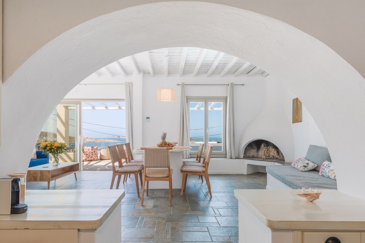 An arched entrance leads to a bright open area with a dining table surrounded by four chairs. A comfortable seating space with a grey sofa is nearby, while large windows showcase views of the sea. Natural light fills the room, enhancing the airy ambiance.