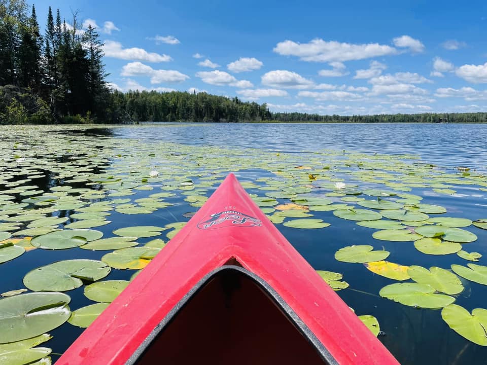 A red kayak is positioned on a calm lake, surrounded by lush green lily pads. The clear blue sky is dotted with fluffy white clouds, and dense trees frame the distant shoreline, creating a serene atmosphere reflective of the natural beauty of the area.