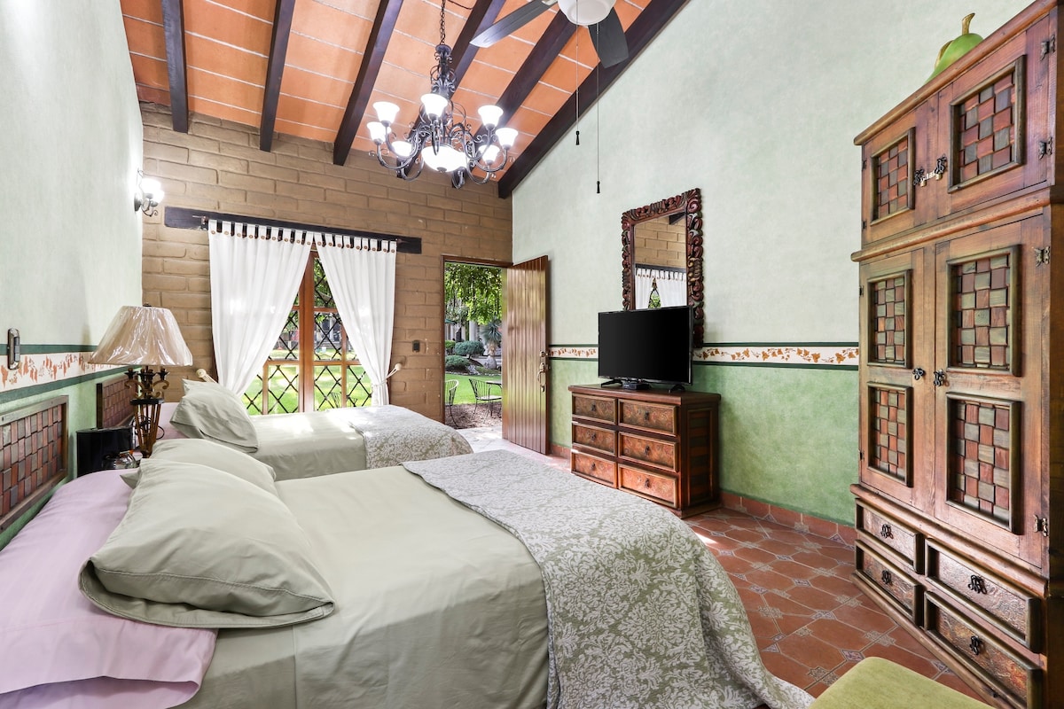 A guest room features two spacious beds with light bedding. Natural light filters through the window adorned with sheer curtains, illuminating the room’s green walls and wooden accents. A wooden cabinet and television provide additional amenities within the rustic decor.