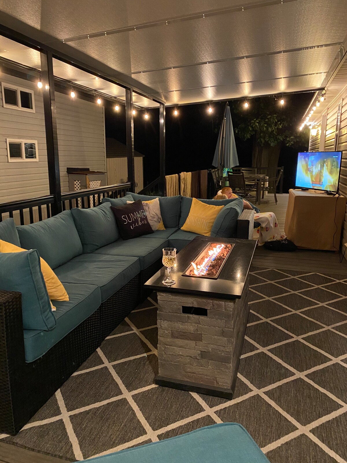 A shaded patio area is featured with a sectional sofa upholstered in blue fabric, complemented by yellow accent pillows. A rectangular fire table in the center provides warmth, while string lights overhead illuminate the space. A large umbrella and entertainment setup are also visible.