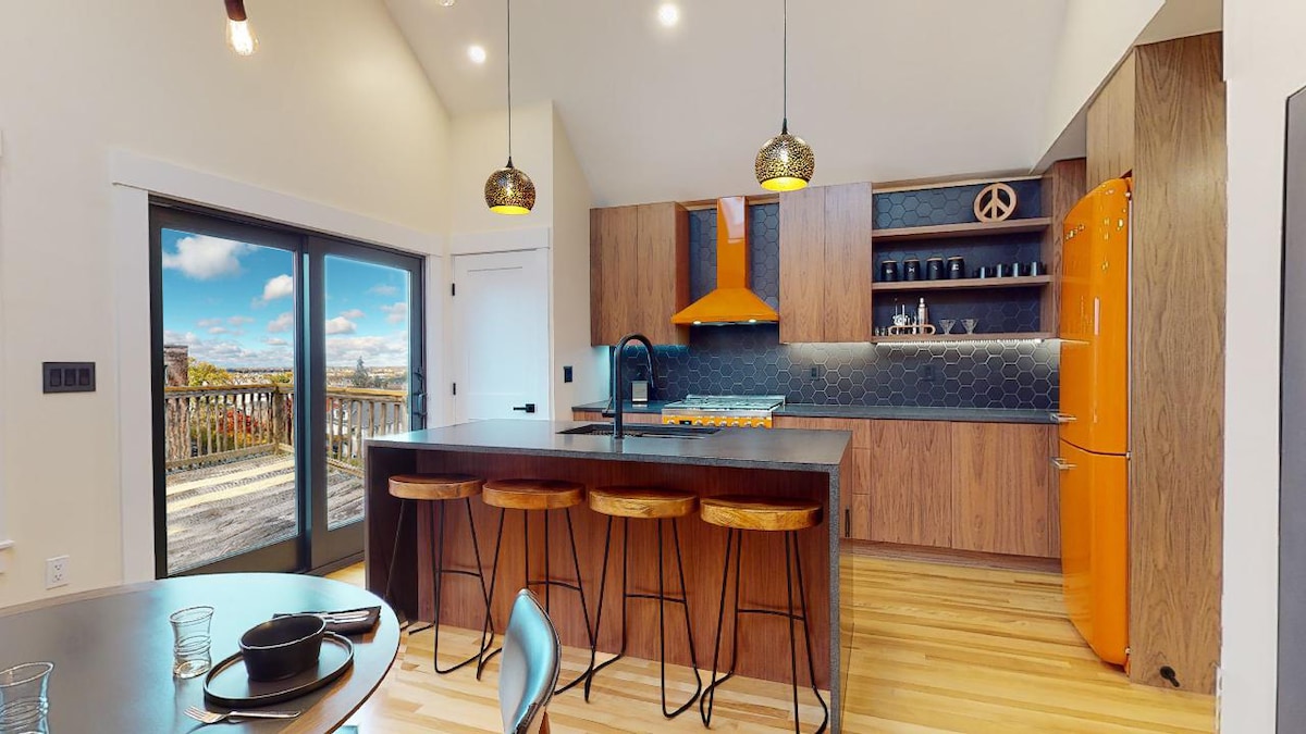 The modern kitchen features a central island with three wooden stools and a bright orange Smeg refrigerator. Dark wood cabinetry complements the sleek black countertops, with a panoramic view visible through sliding glass doors that open to the back deck.