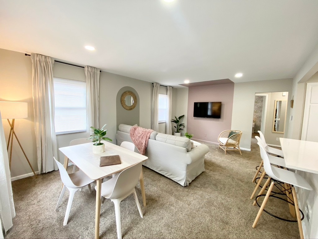 A bright living space showcases a comfortable sectional sofa near a wall-mounted television. A circular mirror reflects natural light, while a dining table for four is positioned beside large windows. Soft carpeting and indoor plants enhance the ambiance, providing a cozy atmosphere.