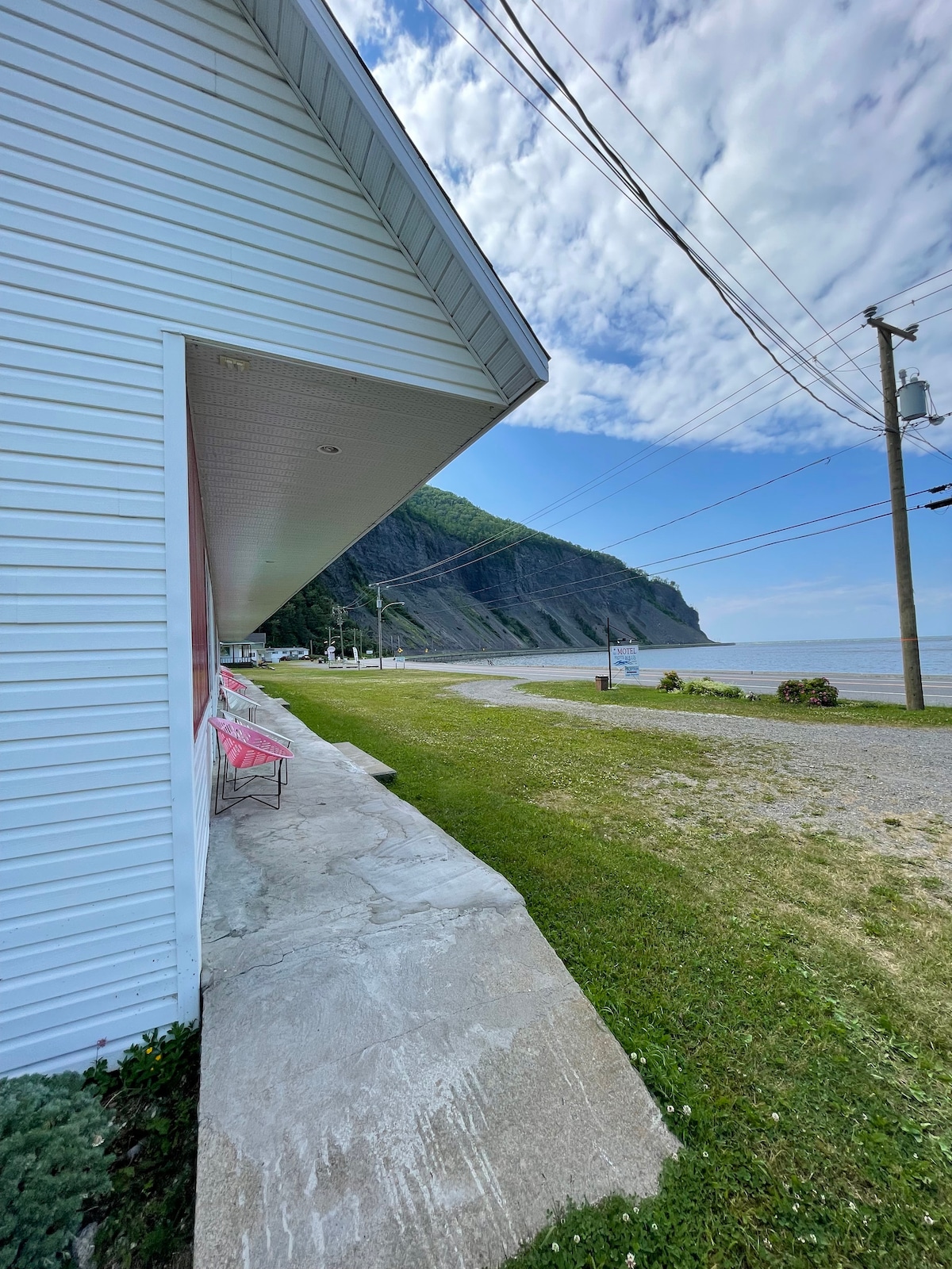 A coastline view is depicted, with a smooth, grassy area leading to a sandy shore. The image captures a portion of a building with light siding, beside which pink chairs are positioned. The backdrop features a rocky cliff and an expansive sky with gentle clouds.