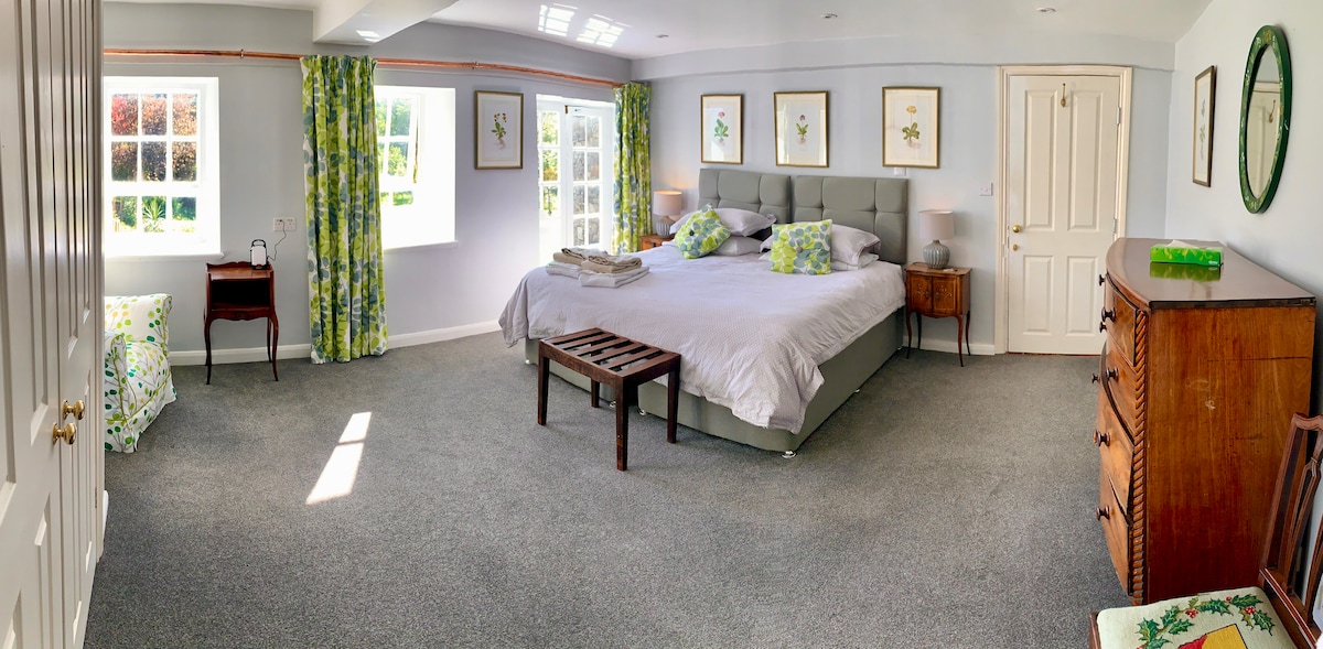 A spacious bedroom is highlighted by large windows that provide natural light. A comfortable king-sized bed, layered with neutral linens and decorative pillows, is positioned centrally. Adjacent seating is available, along with a dresser and side tables, creating a functional retreat.