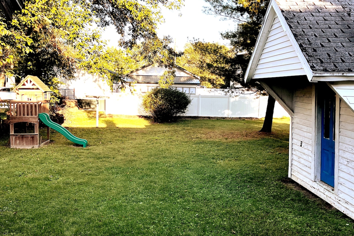 The backyard features a spacious lawn surrounded by trees and a white privacy fence. A playhouse and slide are visible, accompanied by a swing set in a well-shaded area. A small shed is positioned to one side, enhancing the outdoor space.