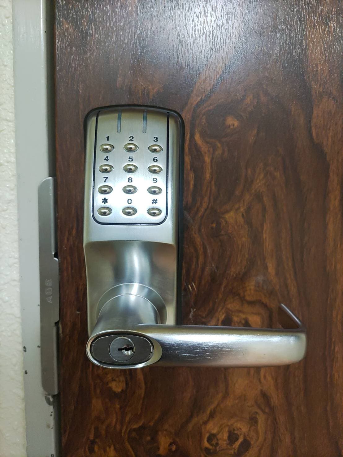 A modern keypad lock is mounted on a wooden door. The lock features a numerical keypad on the upper portion, with a sleek handle below it. The wood grain of the door is visible, displaying a rich texture.