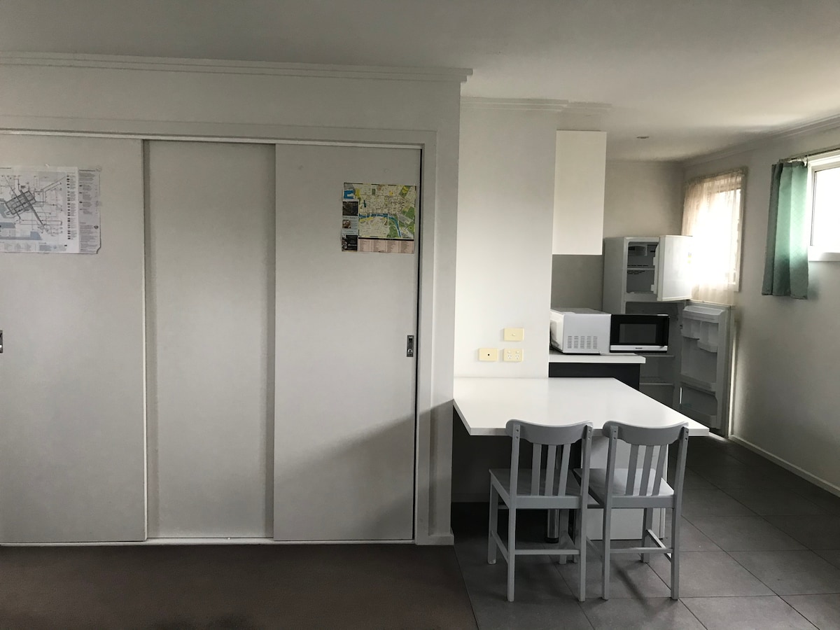 A simple dining area is present, featuring a table and two light-colored chairs. Nearby, basic kitchen appliances, including a microwave and refrigerator, are positioned on a counter. Smooth, neutral-colored walls enhance the studio's spacious feel.