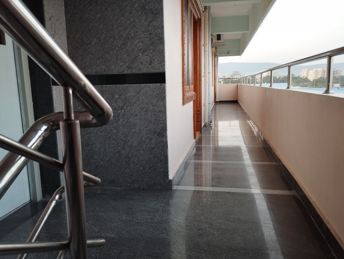 A wide corridor is highlighted by polished stone flooring and decorative wooden doors lining one side. A stainless-steel railing is visible, providing safety along the edge. Natural light filters in from windows, creating a bright and airy atmosphere while offering views of the surroundings.