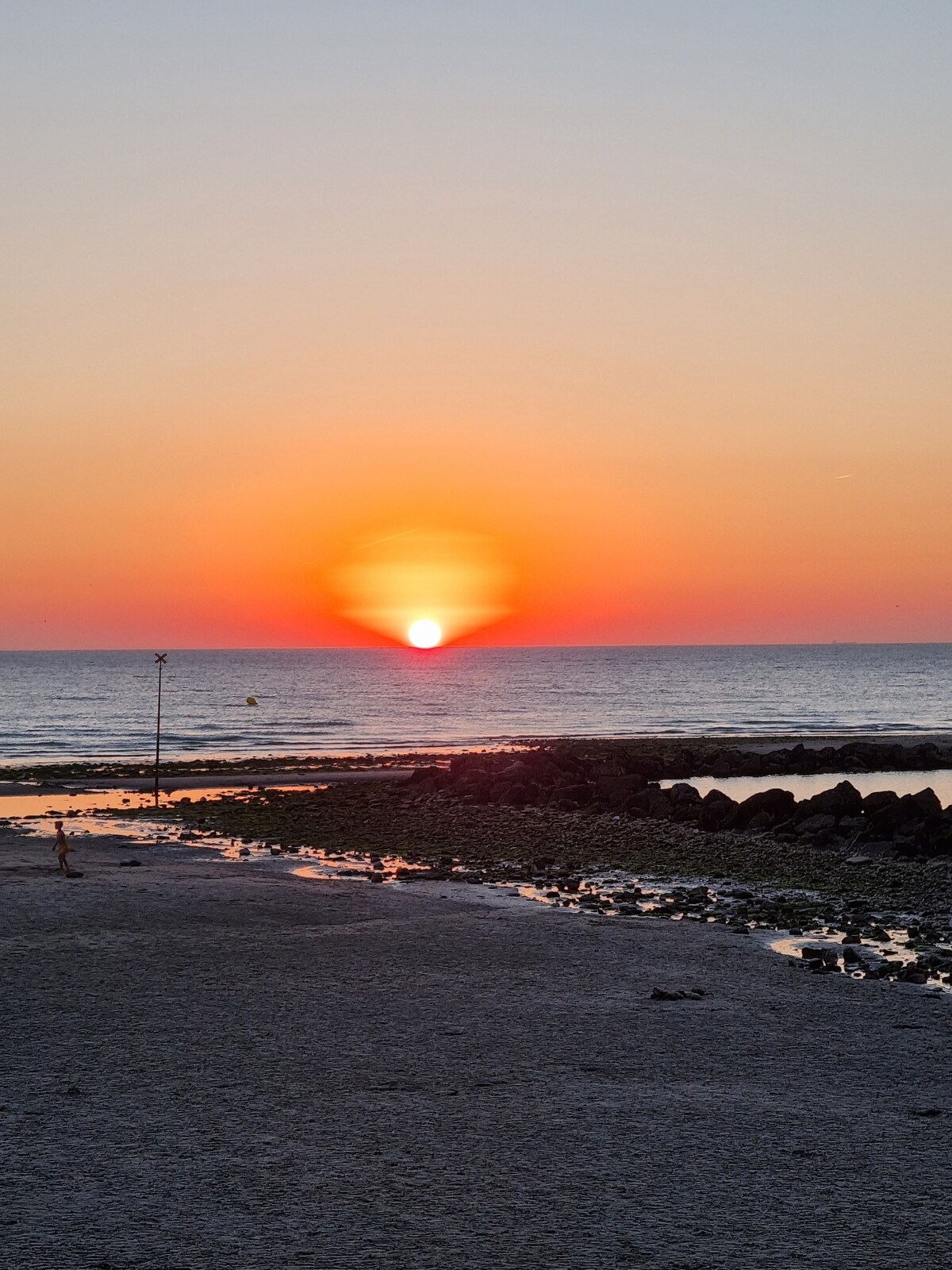 A sunset over the water is depicted, with vibrant orange and pink hues blending into the sky. The sun is halfway above the horizon, reflecting off the calm sea. The foreground features a sandy beach with scattered rocks and tidal pools, capturing the tranquility of the scene.