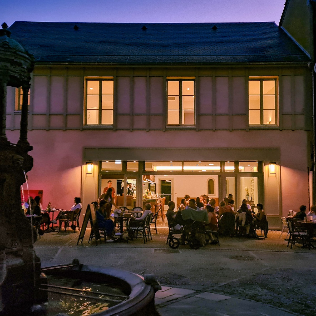 An outdoor dining area is visible, featuring tables arranged in groups with diners enjoying their meals. Soft lighting from the restaurant's interior illuminates the scene. The historic building's large windows and fountain contribute to a welcoming atmosphere.