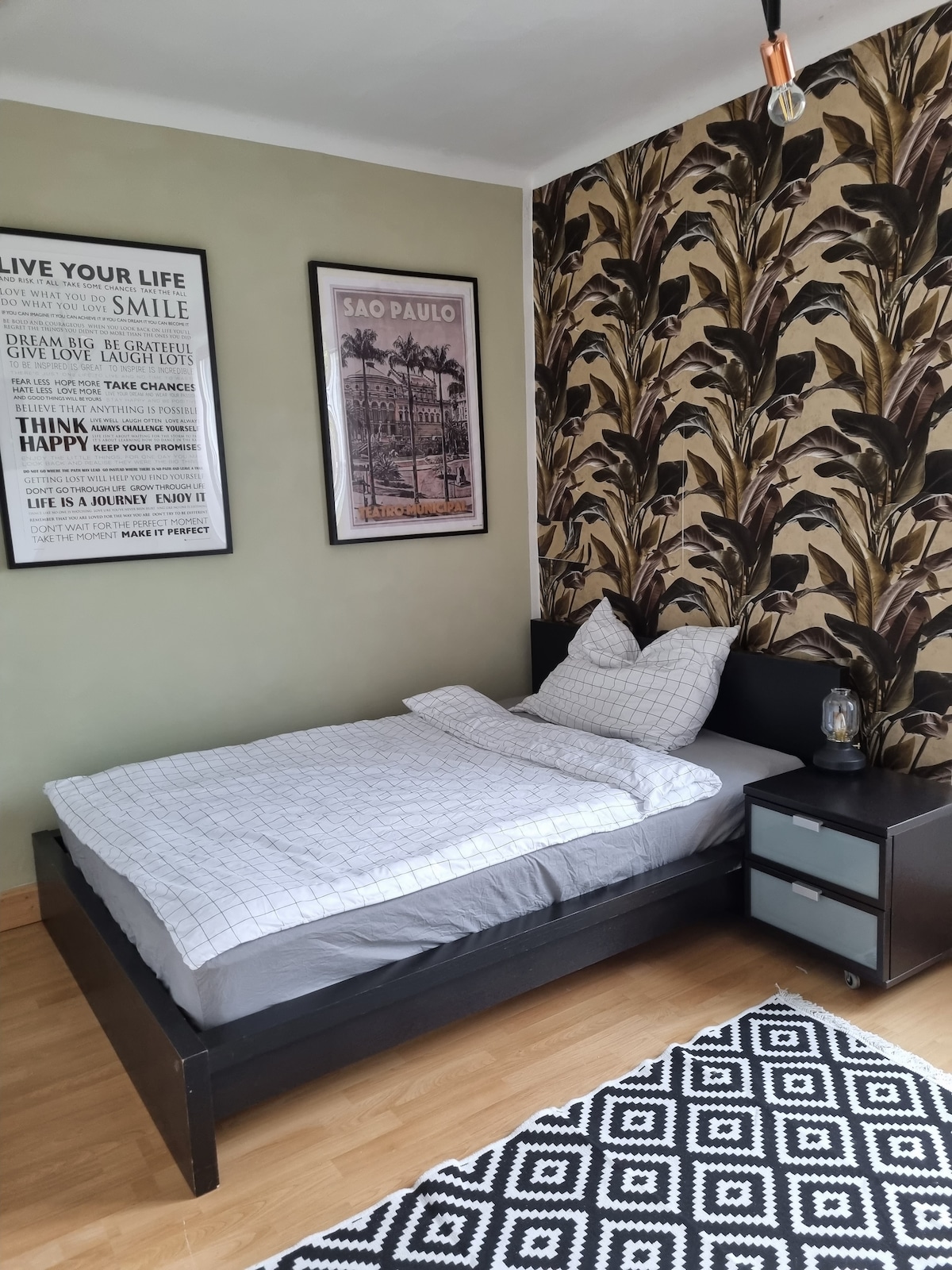 A cozy sleeping area is highlighted by a spacious bed with neutral bedding. The wall is adorned with a tropical leaf pattern, complemented by framed artwork. A nightstand with a drawer sits beside the bed, and a geometric rug adds texture to the wooden floor.