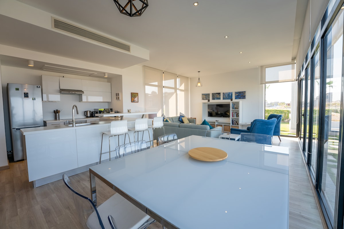 An open-concept living area is highlighted by large glass doors that invite natural light. A modern kitchen with sleek cabinetry and bar seating adjoins a dining table. Comfortable seating is arranged in the lounge area, complemented by accent decor and views of the outdoors.