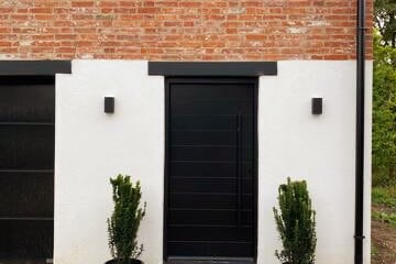 The entrance features a sleek black door framed by a bright white exterior wall. Two slender potted plants are positioned on either side of the door. The textured red brick wall above adds a rustic detail to the contemporary design.