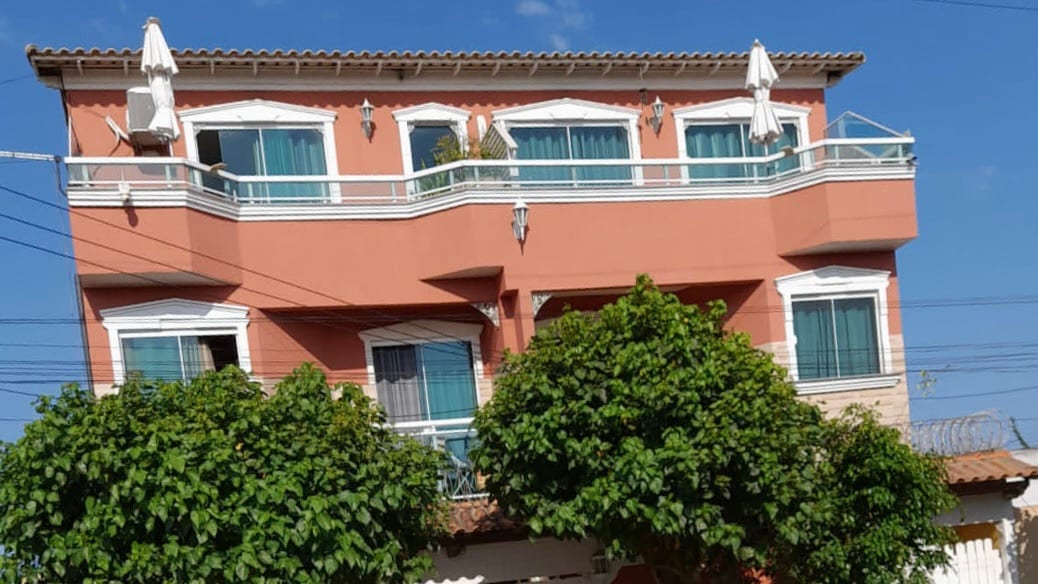 A two-story building features a peach-colored facade with multiple balconies adorned with awnings. Large windows are framed with green curtains, offering a bright and airy appearance. Lush trees are positioned in the foreground, providing greenery and a sense of tranquility.