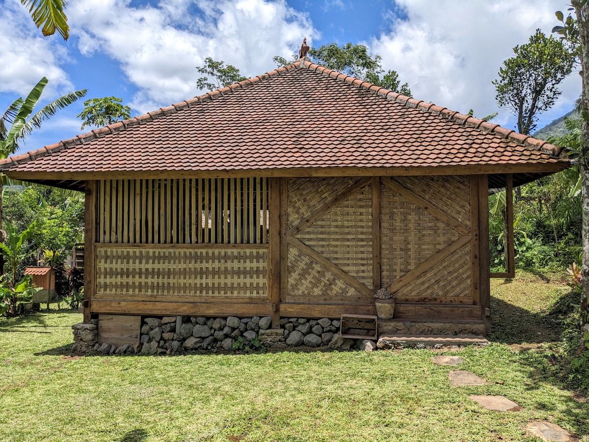 The exterior of a traditional wooden cottage is showcased, featuring a sloped roof with clay tiles. Bamboo panels and wooden accents create a rustic appearance. A well-maintained garden surrounds the structure, with stone pathways leading to the entrance, framed by lush greenery.