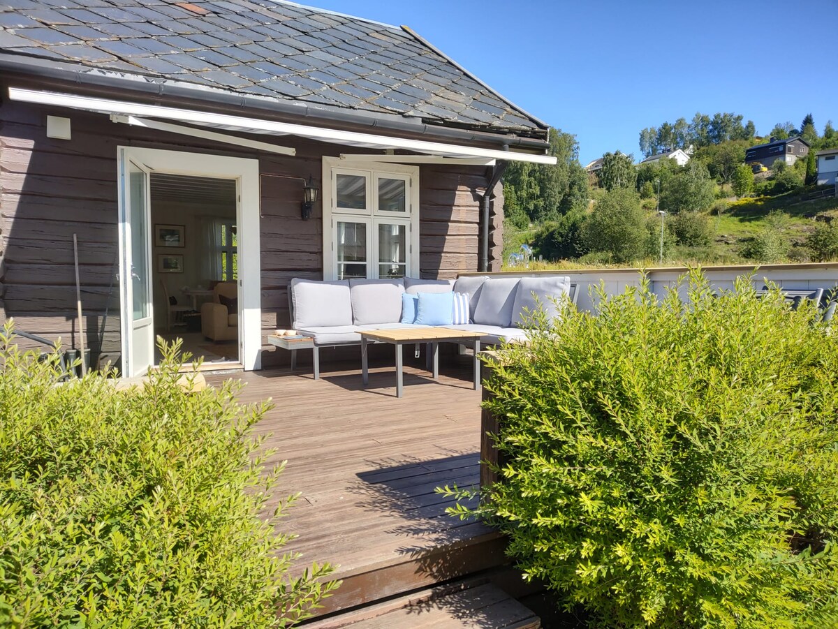 A wooden deck area is presented, featuring a spacious outdoor seating arrangement with soft cushions and a central table. The entryway to the interior is visible through an open door, and greenery surrounds the space, complemented by a panoramic view of the surrounding hills.