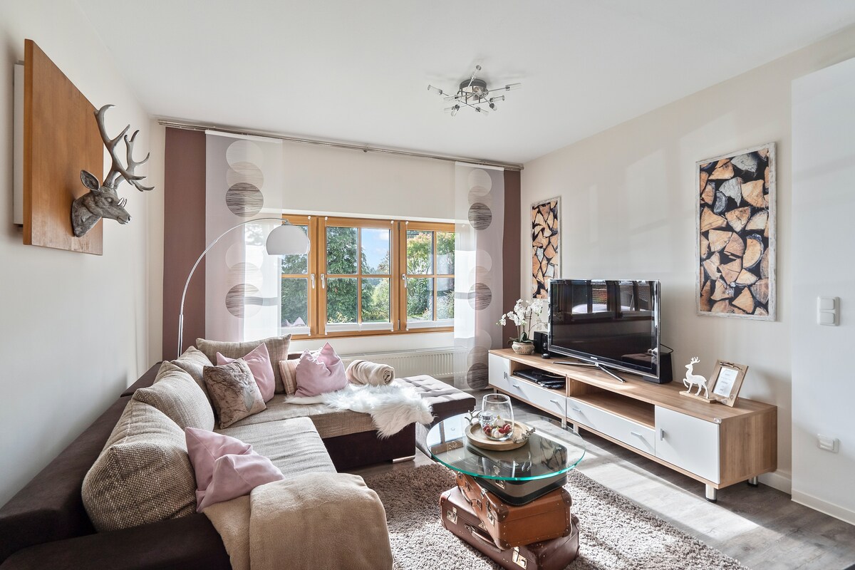 A spacious living area features a sectional sofa adorned with cozy throws and cushions. Large windows allow natural light to flood the room, while a glass coffee table sits in the center. Decorative wall art and a mounted deer head add character to the neutral decor.