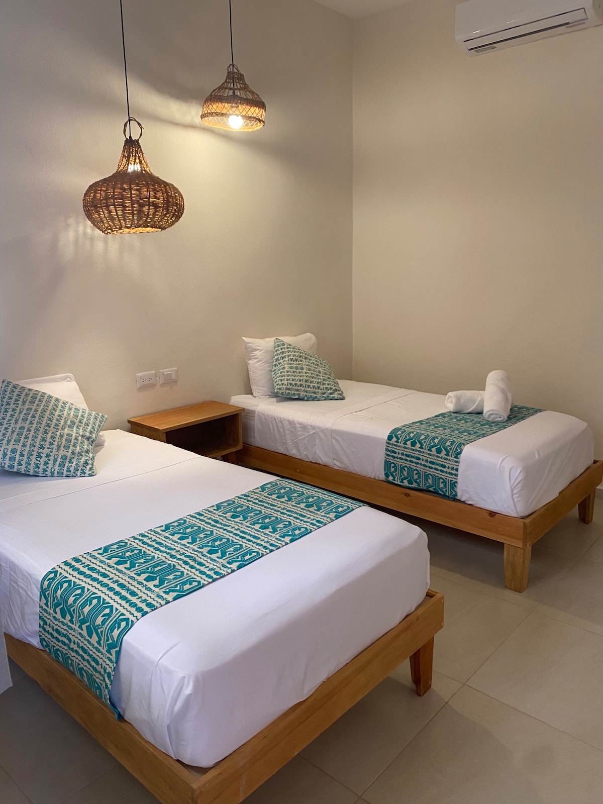 The room features two twin beds with decorative turquoise throws, complemented by rustic wooden bed frames. Each bed is accompanied by a small nightstand. Soft lighting is provided by hanging woven pendant lamps, creating a calm and bright environment.