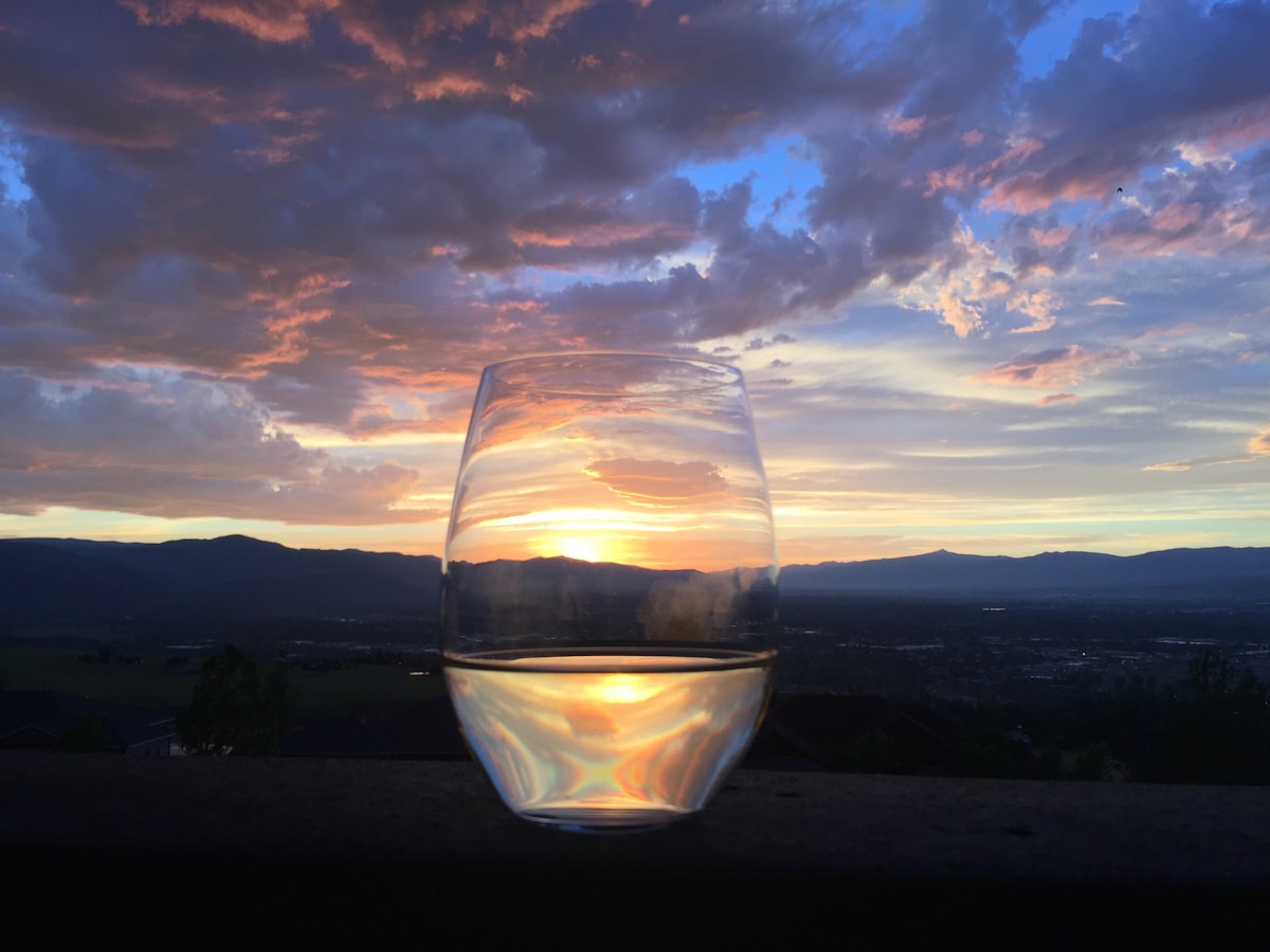 A clear glass sits on a surface, reflecting the warm hues of a sunset. The sky displays a blend of orange, pink, and purple tones, with distant mountains lining the horizon under the colorful cloud formations.