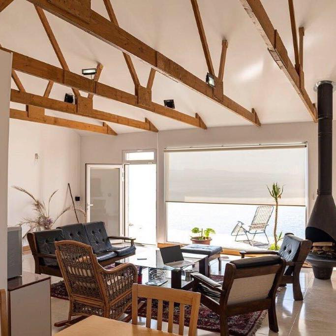 A spacious living area features high wooden beams and large windows that allow natural light. Comfortable seating arrangements include dark wooden chairs and a low table, complemented by a rug. A sleek fireplace adds a modern touch to the inviting space.