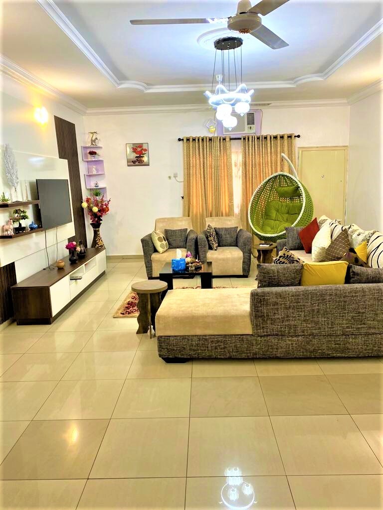 A spacious living room is featured, furnished with a large sectional sofa and a hanging chair. Decorative throw pillows add color to the seating area, while a coffee table is positioned at the center. Soft lighting and a ceiling fan enhance the comfortable ambiance.