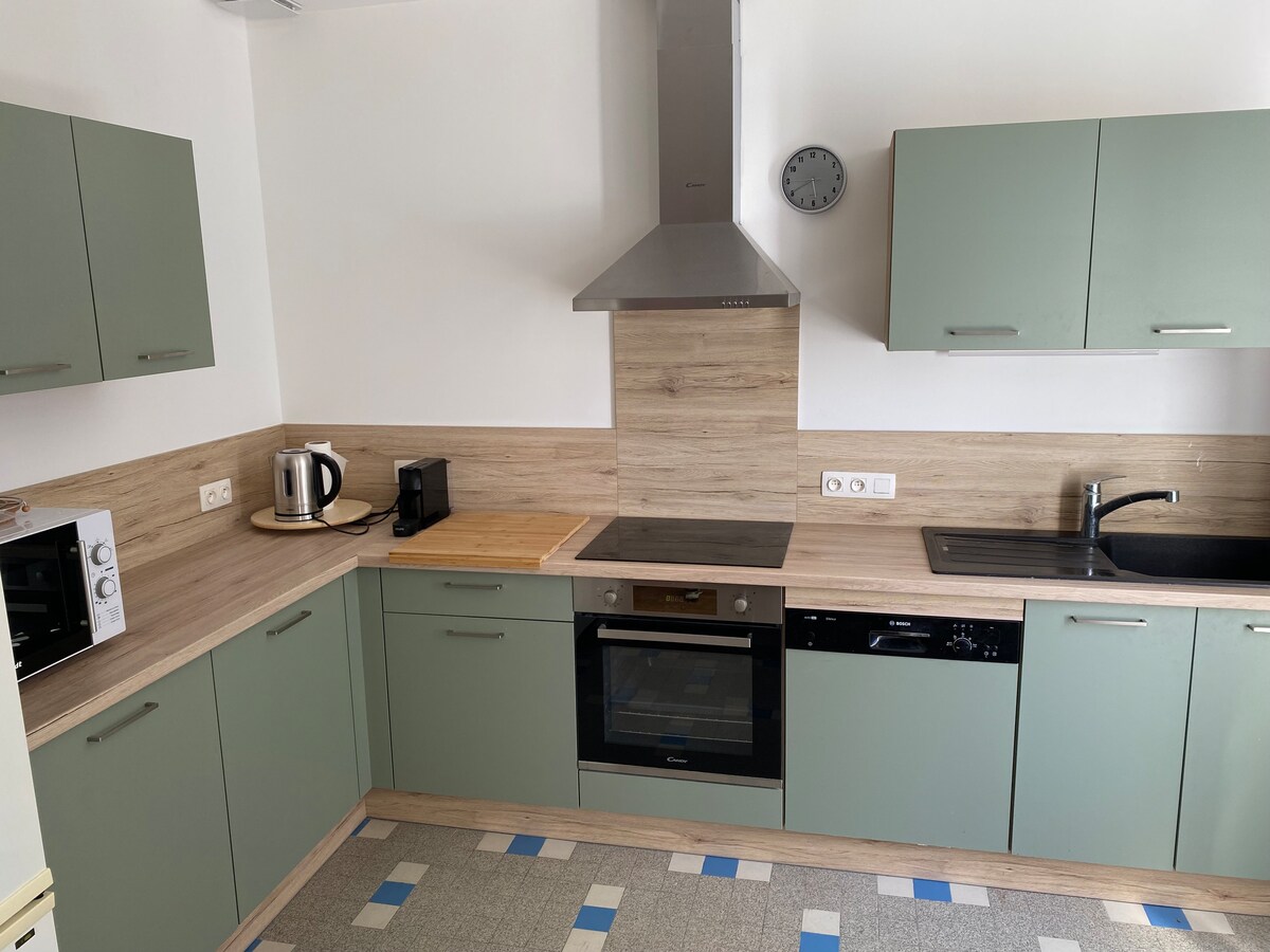 The kitchen showcases modern cabinetry in muted green tones, complemented by a neutral countertop. A stainless steel oven and microwave are visible, along with a kettle and coffee machine. The space is well-lit, featuring a hood above the stove and a clock mounted on the wall.