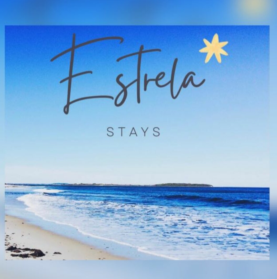 A serene beach scene features a gentle shore where soft waves meet the sandy coastline. The clear blue sky stretches overhead, creating a tranquil atmosphere. The words 'Estrela Stays' are artistically displayed, adding a touch of charm to the image.