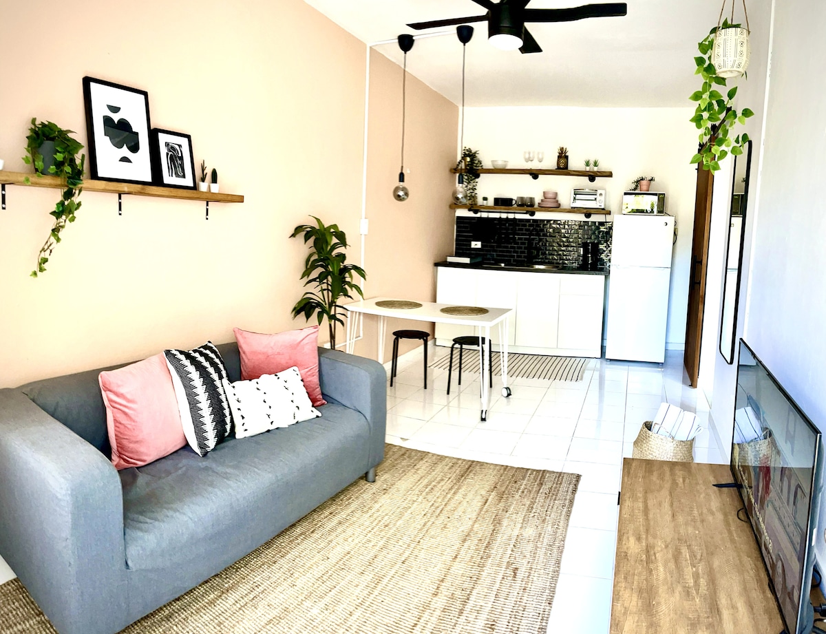 A modern living space features a gray sofa with decorative pillows, paired with a light wooden coffee table. A small dining area with a white table and chairs is positioned in front of the kitchenette, which is equipped with sleek black cabinetry and white appliances. Natural light fills the room through a large window.
